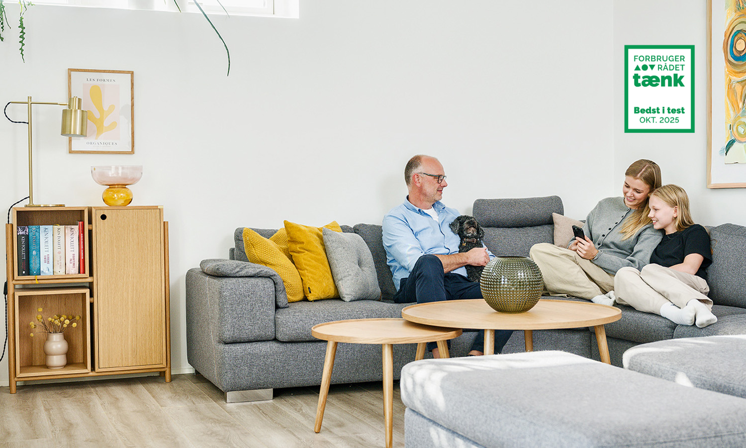 Tre personer og en hund sidder på en grå sofa i et stue. En vægdekoration med tekst er på væggen.